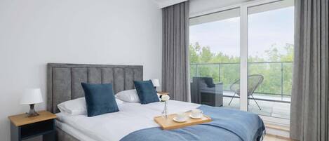Une chambre d’hôtel moderne avec un grand lit, une table de chevet, une petite table sur laquelle se trouve un plateau avec des articles pour le petit-déjeuner, et un balcon avec une chaise.
