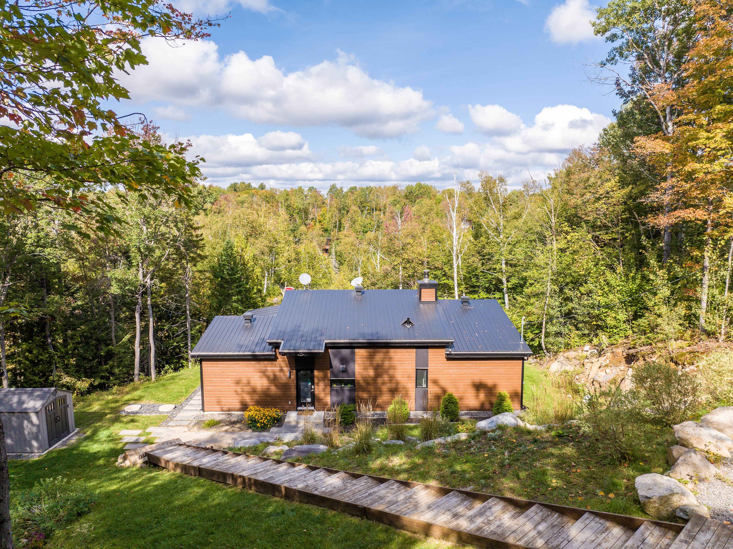 A modern house with a dark roof, surrounded by a well-maintained garden and a forested area.
