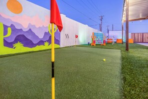 Un campo de mini golf con césped artificial, un mural colorido y una pequeña zona de descanso con cojines anaranjados.