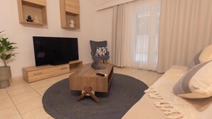 Ett modernt vardagsrum med en platt-TV, trähyllor, en grå fåtölj, en soffa och ett stort fönster med gardiner.