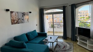 Een moderne woonkamer met een blauwgroene bank, een salontafel en een flatscreen-tv. Op de muur hangen abstracte bladschilderijen en er is uitzicht op een balkon met gebouwen op de achtergrond.