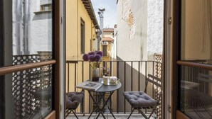 Un balcón con una mesa, dos sillas y un jarrón de flores moradas.