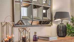 Una mesita auxiliar de madera con una lámpara, dos velas, un libro titulado 'PHOTOGRAPHY' y un espejo decorativo con diseños geométricos.