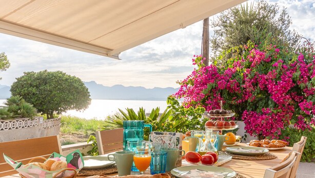 An outdoor dining table with a view of the sea, surrounded by flowers and various breakfast items.