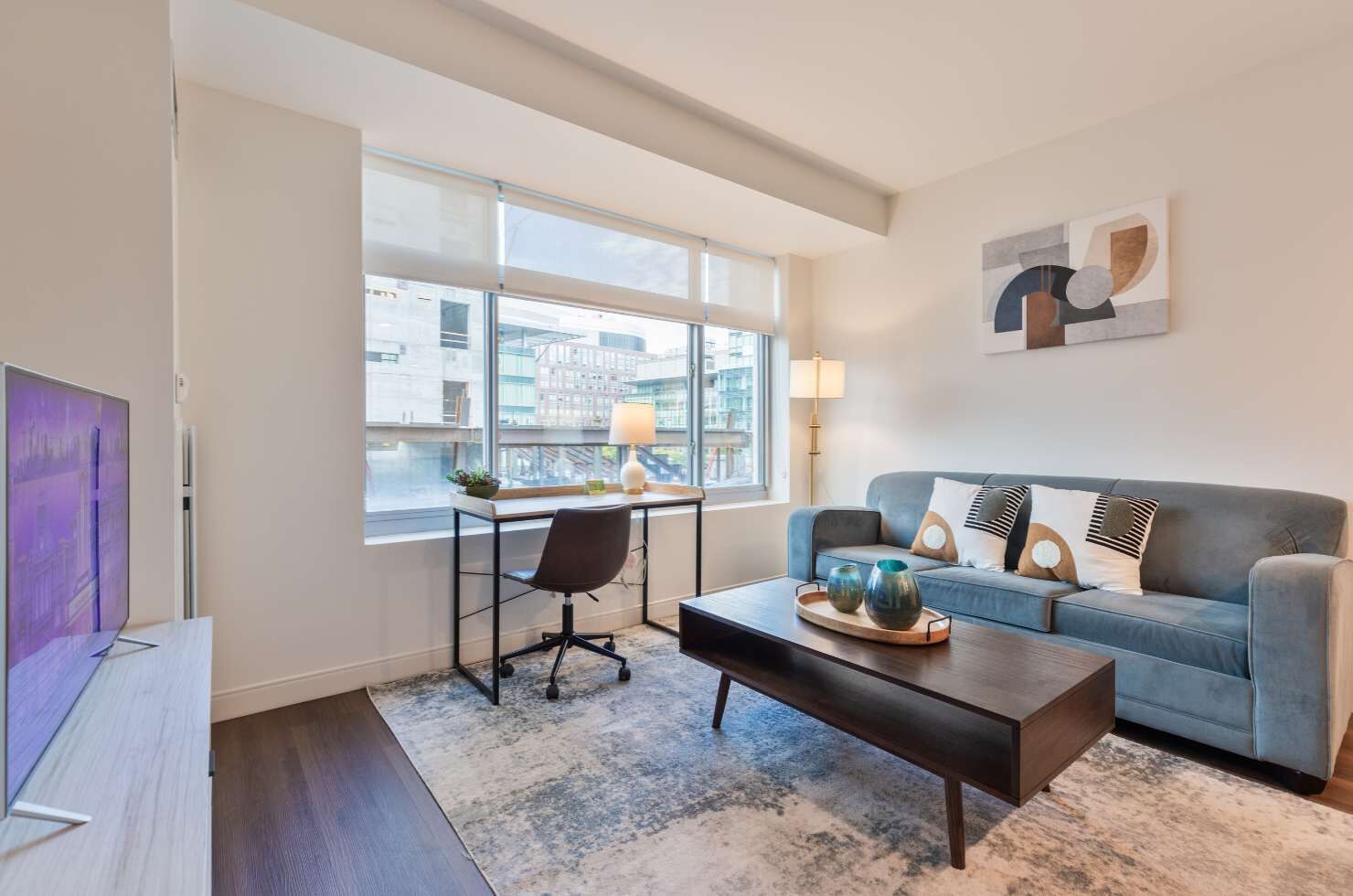 A modern living room with a sofa, coffee table, and desk. There is a large window with a view of buildings outside.