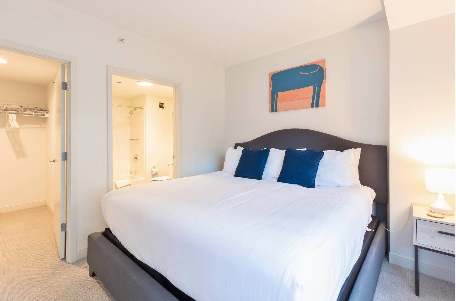 A hotel room with a large bed, a bedside table with a lamp, a bathroom with a shower and toilet, and a closet with hanging clothes.