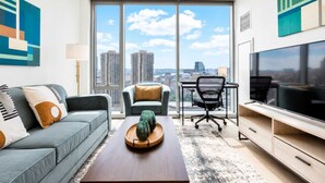 A modern living room with a sofa, coffee table, and a desk with a chair, large TV, and a city view through floor-to-ceiling windows. - Luxury Apt Near TD Garden (Boston)