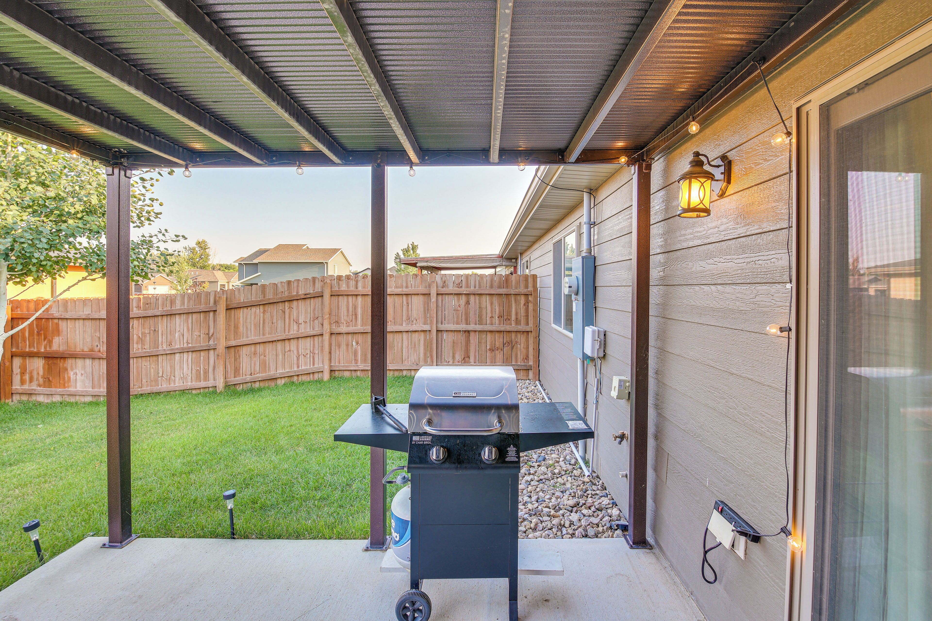 A backyard with a barbecue grill under a covered patio, a wooden fence, and a house in the background.