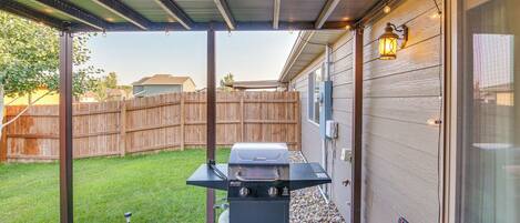 A backyard with a barbecue grill under a covered patio, a wooden fence, and a house in the background.