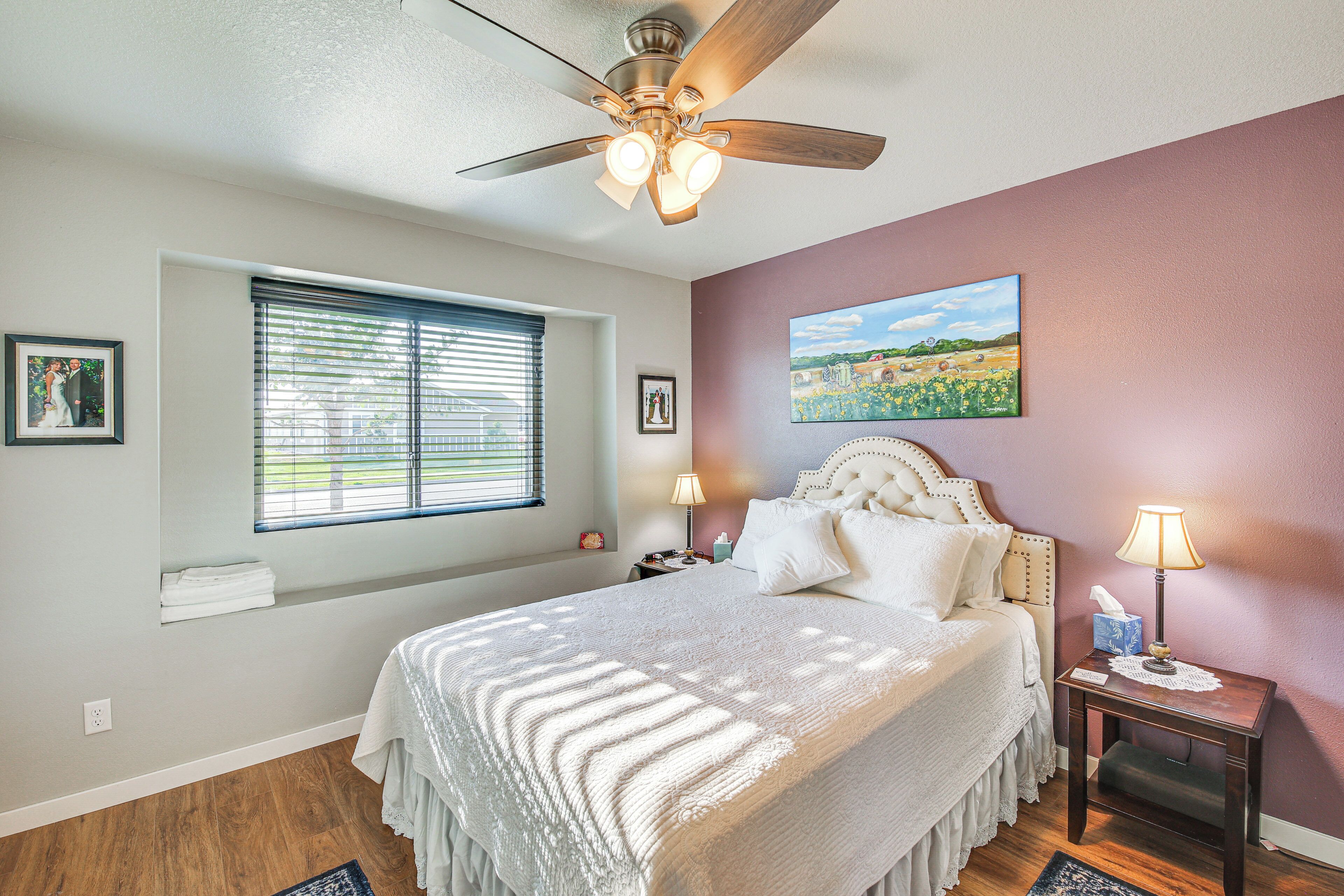 Une chambre à coucher comprenant un lit, une table de chevet, une fenêtre avec des stores, un ventilateur de plafond et un tableau accroché au mur.