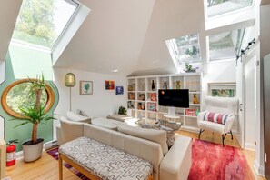 A modern living room with a sofa, armchair, and a TV. There are bookshelves, a plant, and a fire extinguisher.