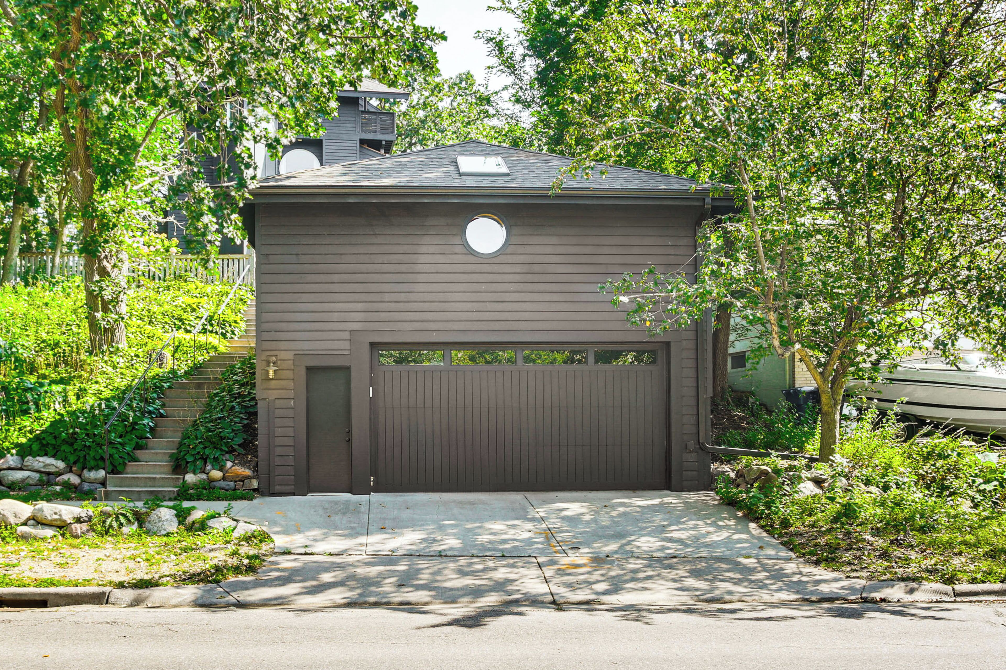 A two-story house with a garage, surrounded by lush greenery and a stone staircase leading to the entrance.