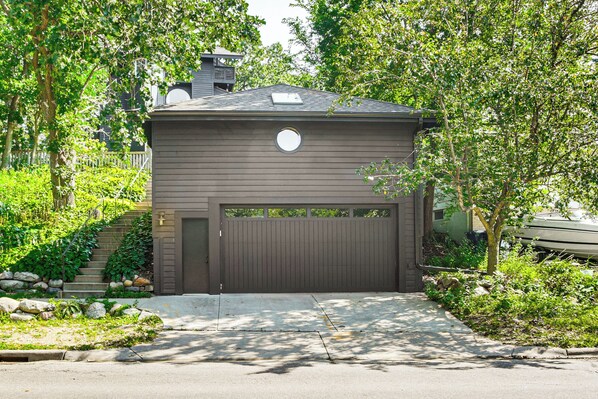A two-story house with a garage, surrounded by lush greenery and a stone staircase leading to the entrance.