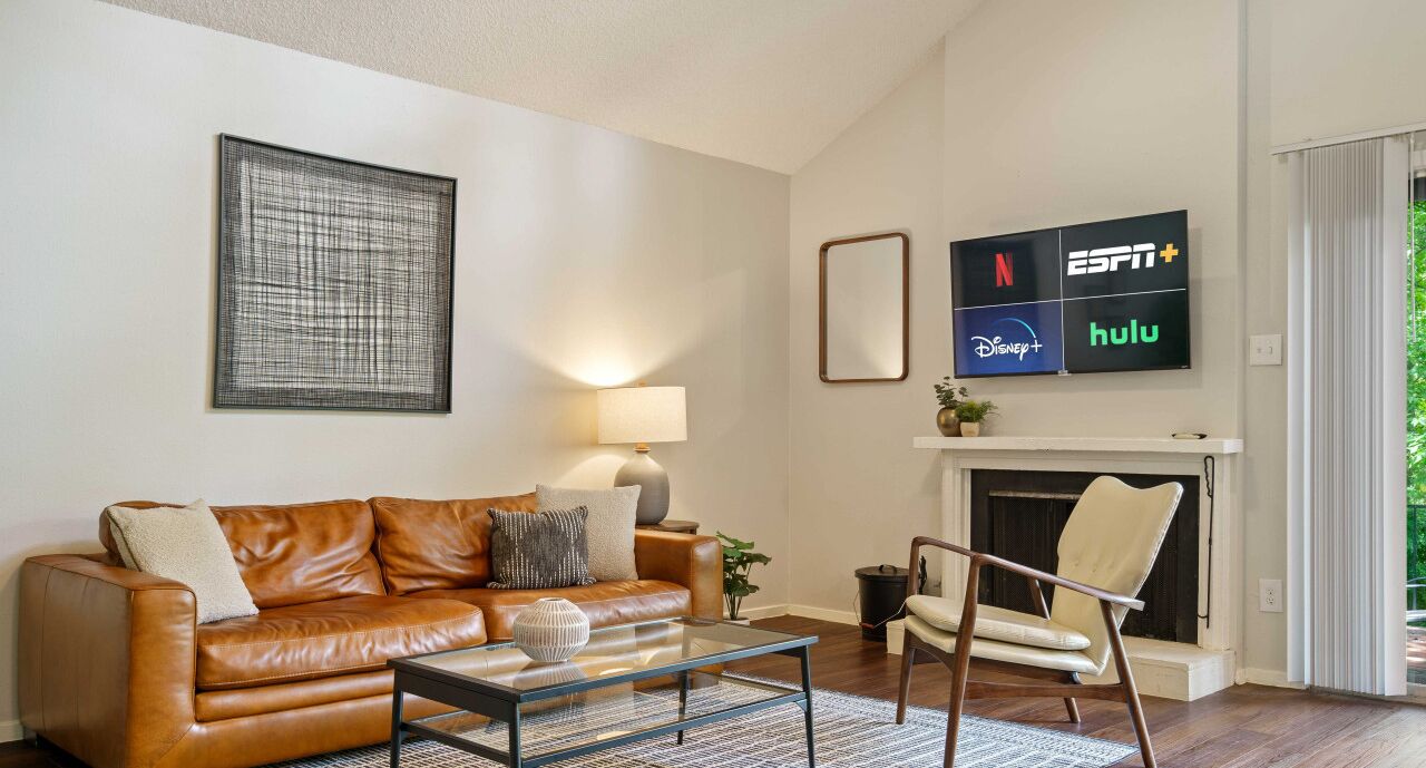 A living room with a brown leather sofa, a glass coffee table, a white armchair, a fireplace, and a wall-mounted TV displaying streaming services.