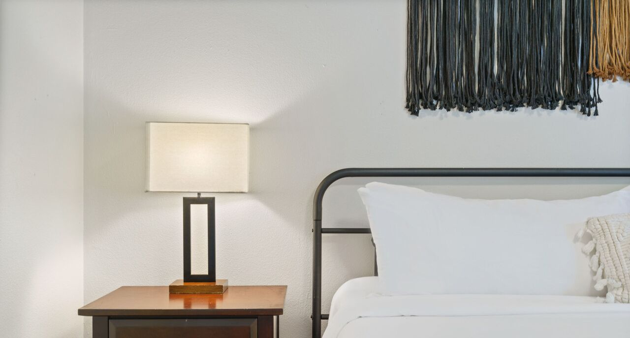 A hotel room with a bed, a nightstand with a lamp, and a wall with a hanging textile.