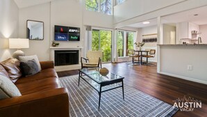 Un salón espacioso con chimenea, una mesa de centro de vidrio y un sofá de cuero. Hay un televisor montado en la pared mostrando servicios de streaming.