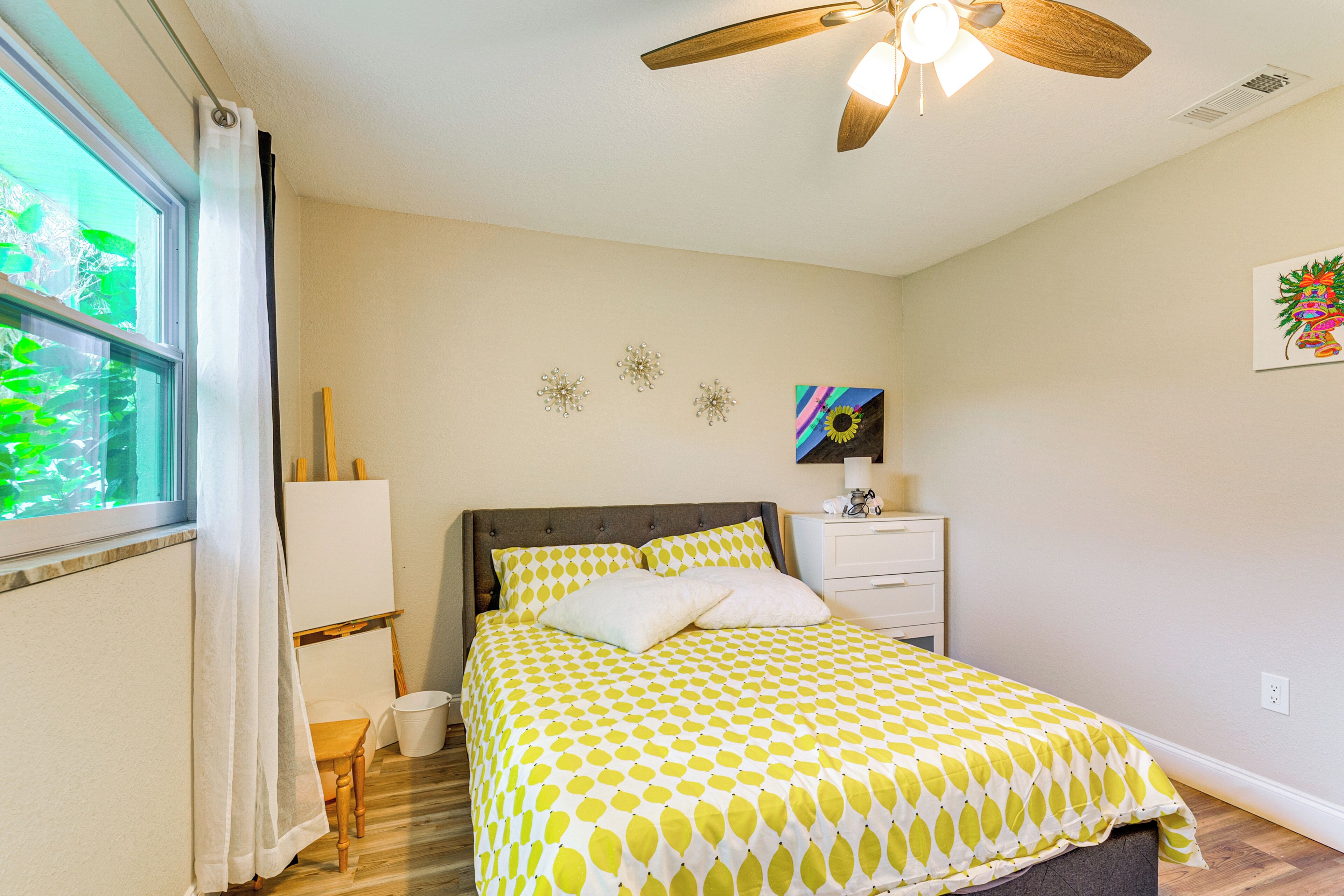 Une chambre à coucher comprenant un lit, un ventilateur de plafond, une table de chevet, une fenêtre avec des rideaux et des éléments de décoration murale.