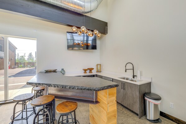 A modern kitchen with a large island, bar stools, a stainless steel sink, and a TV mounted above the island. - Ski & Stroll: Condo Near Whitefish Resort (Whitefish)