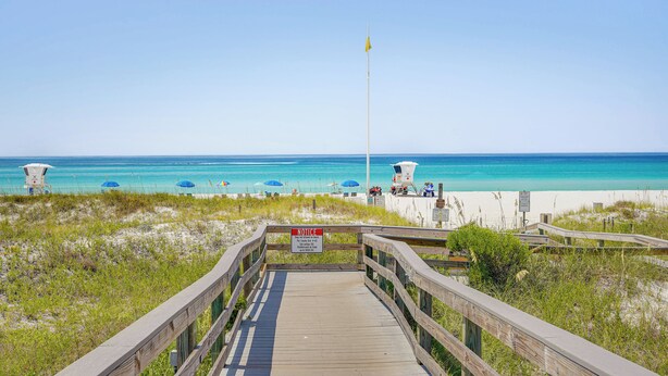A wooden boardwalk leading to a beach with clear blue water, a lifeguard tower, and beach umbrellas. - Panama City Home w/ Fire Pit - Walk to Beaches! (Panama City Beach)