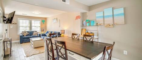 A spacious living room with a large wooden dining table surrounded by chairs, a blue sofa with patterned cushions, and a kitchen area with a wooden countertop and various items on the shelves.