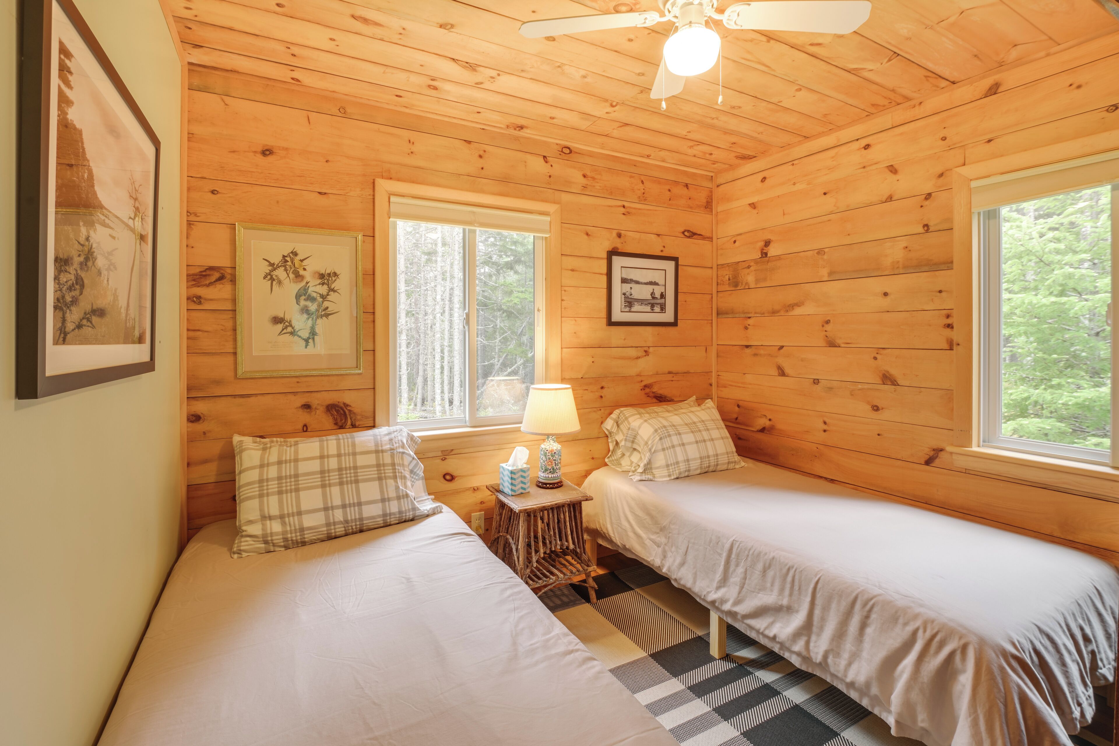 A cozy cabin room with two beds, wooden walls, a ceiling fan, and framed artwork.