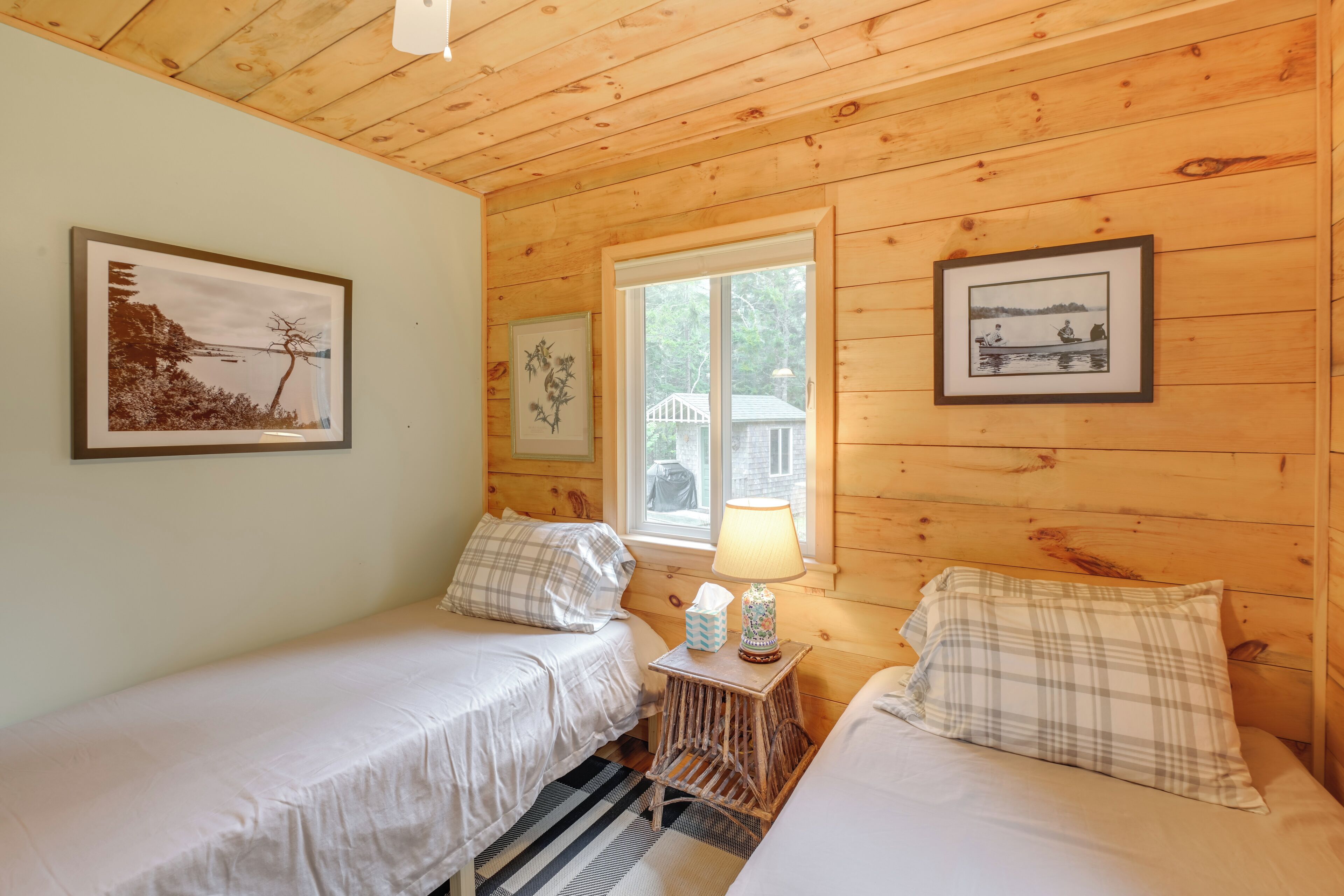A cozy cabin room with two beds, wooden walls, a window with a view, and framed pictures on the walls.