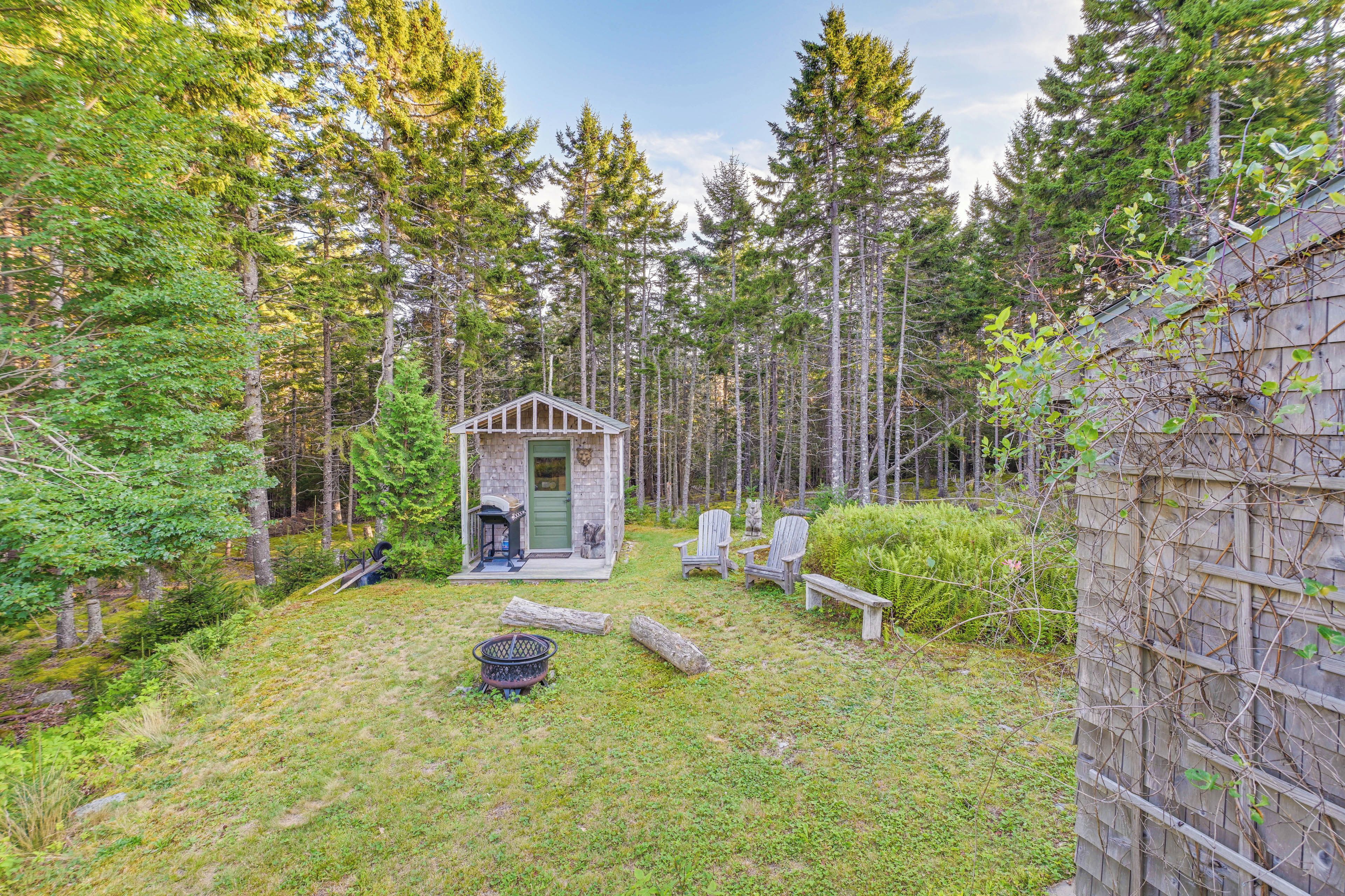 A small wooden cabin with a porch, surrounded by tall trees and a grassy yard.