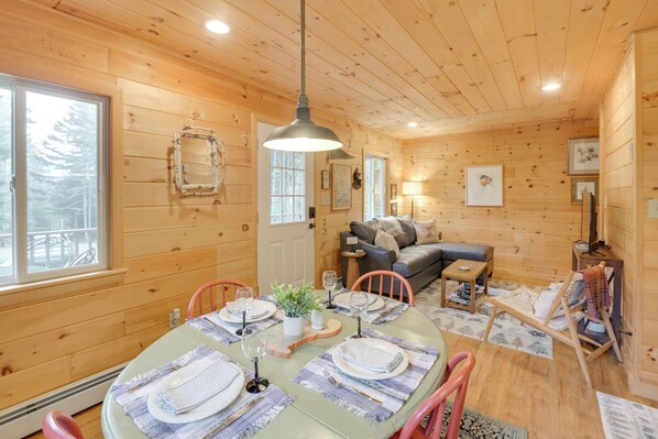 A cozy cabin interior with a dining area set for four, a sofa, and a small table with a lamp. - Cozy Acadia Area Cabin, Walk to Beach & Lighthouse (Prospect Harbor)