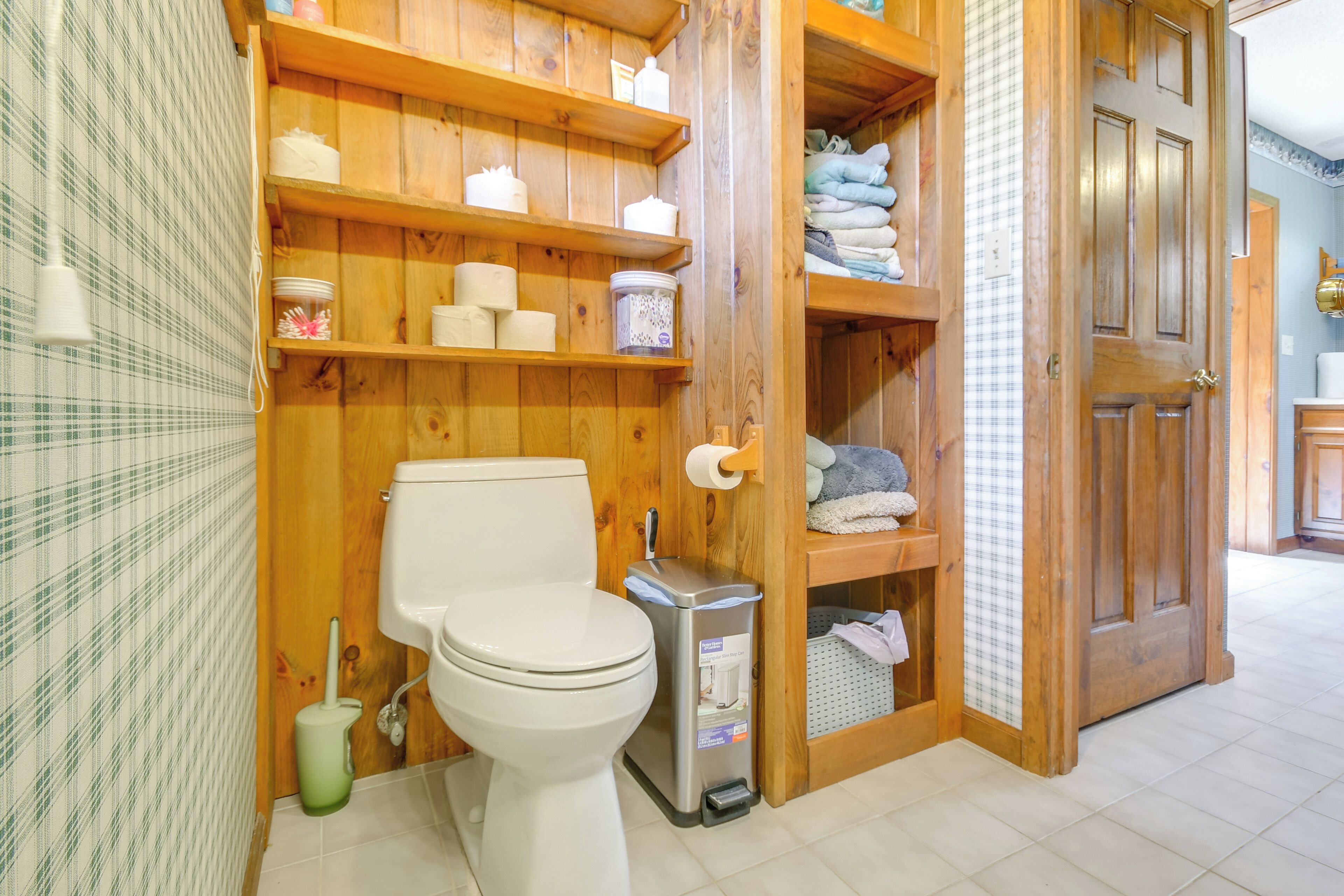 A wooden bathroom with a toilet, shelves with toiletries, and a trash bin.