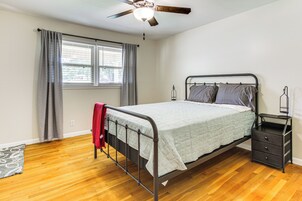 Un dormitorio con estructura de cama metálica, una mesita de noche con cajones, ventilador de techo y una ventana con cortinas.