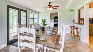 A dining room with a wooden table set for six, surrounded by white chairs. There is a ceiling fan above the table, and a kitchen area is visible in the background. - Spacious Micanopy Farmhouse w/ Luxurious Pool! (Micanopy)