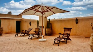 A patio with four wooden lounge chairs, a large umbrella, and potted plants against a textured wall under a clear sky. - Riad (Marrakech)