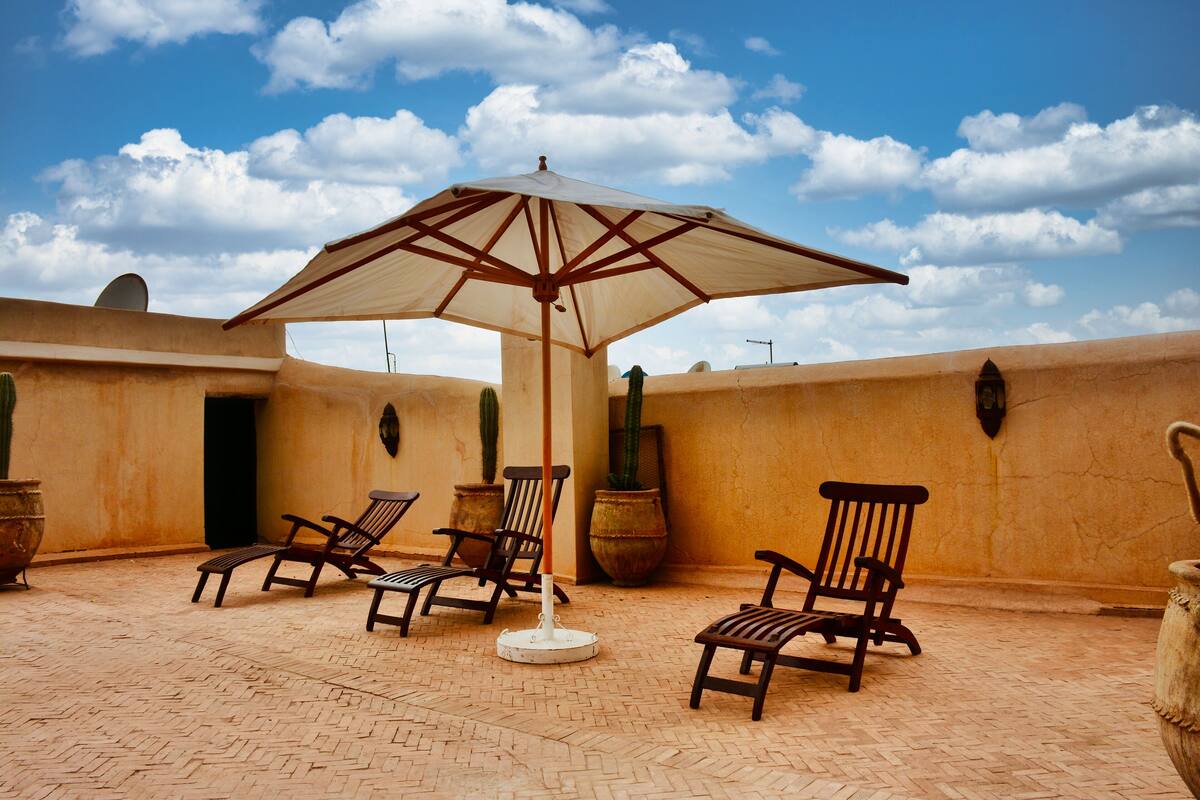 A patio with four wooden lounge chairs, a large umbrella, and potted plants against a textured wall under a clear sky.