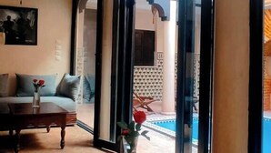 A room with a wooden table, a sofa, and a view of a swimming pool through an open doorway. - Riad (Marrakech)