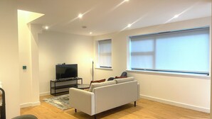 A modern living room with a flat-screen TV on a stand, a grey sofa, a coffee table, and a large window with blinds. - Cosy Modern Townhouse, 5mins to Tube, 02, Excel (London)