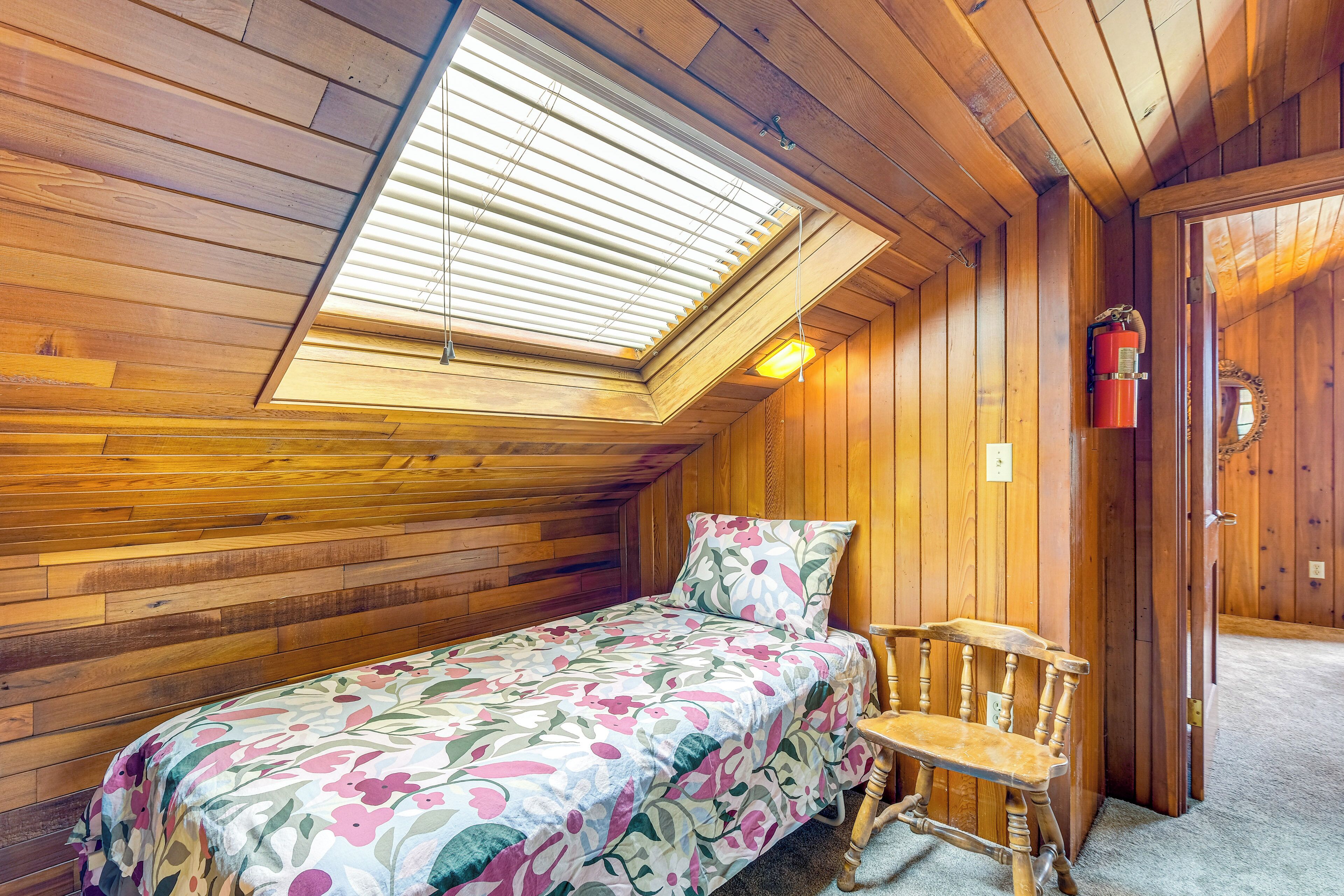 Une petite pièce agréable, dotée d’un plafond en pente, d’un lit recouvert d’une courtepointe à motifs floraux, d’un banc en bois et d’un extincteur fixé au mur.