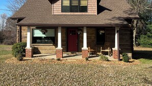 A two-story log cabin with a red door, a covered porch with chairs, and a small front yard with scattered leaves. -  Quaint,newly remodeled country cabin in the heart of Bourbon Country. (Raywick)