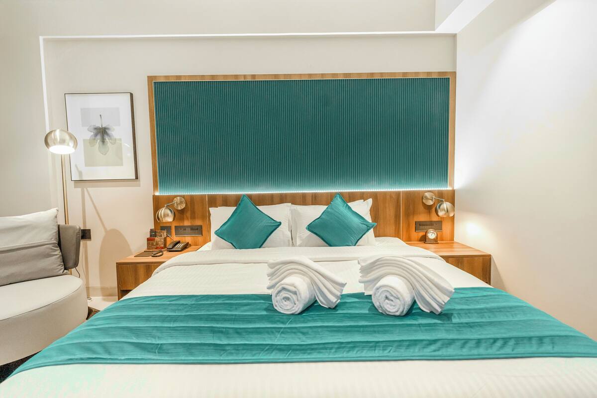 A hotel room with a neatly made bed, a wooden headboard, teal pillows, and a teal bedspread.