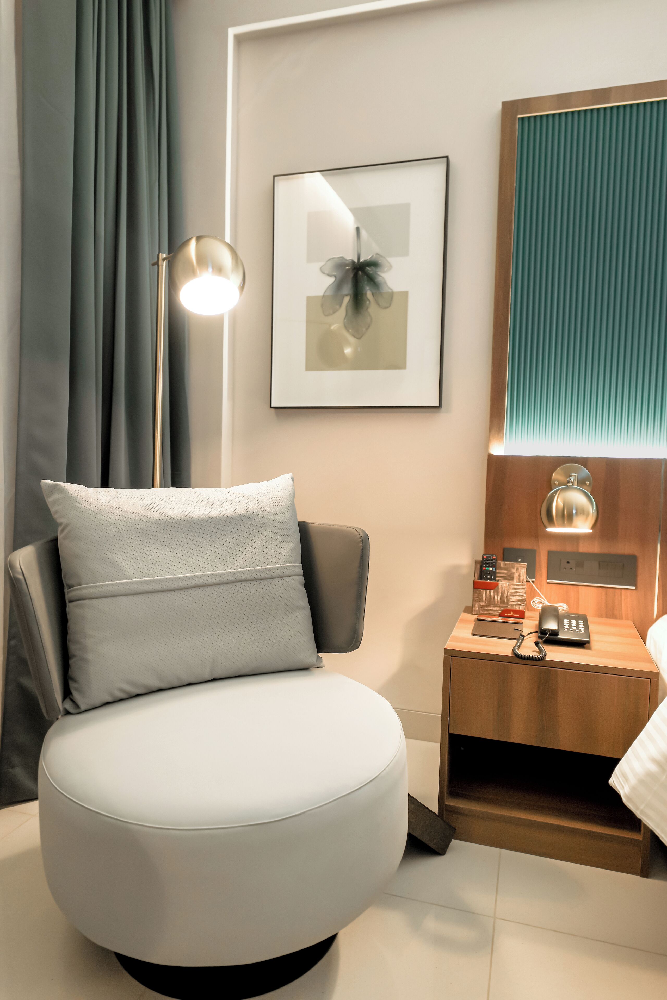 Quarto de hotel moderno com uma cadeira branca arredondada, uma mesa de cabeceira com abajur, um quadro na parede e uma janela com persianas.