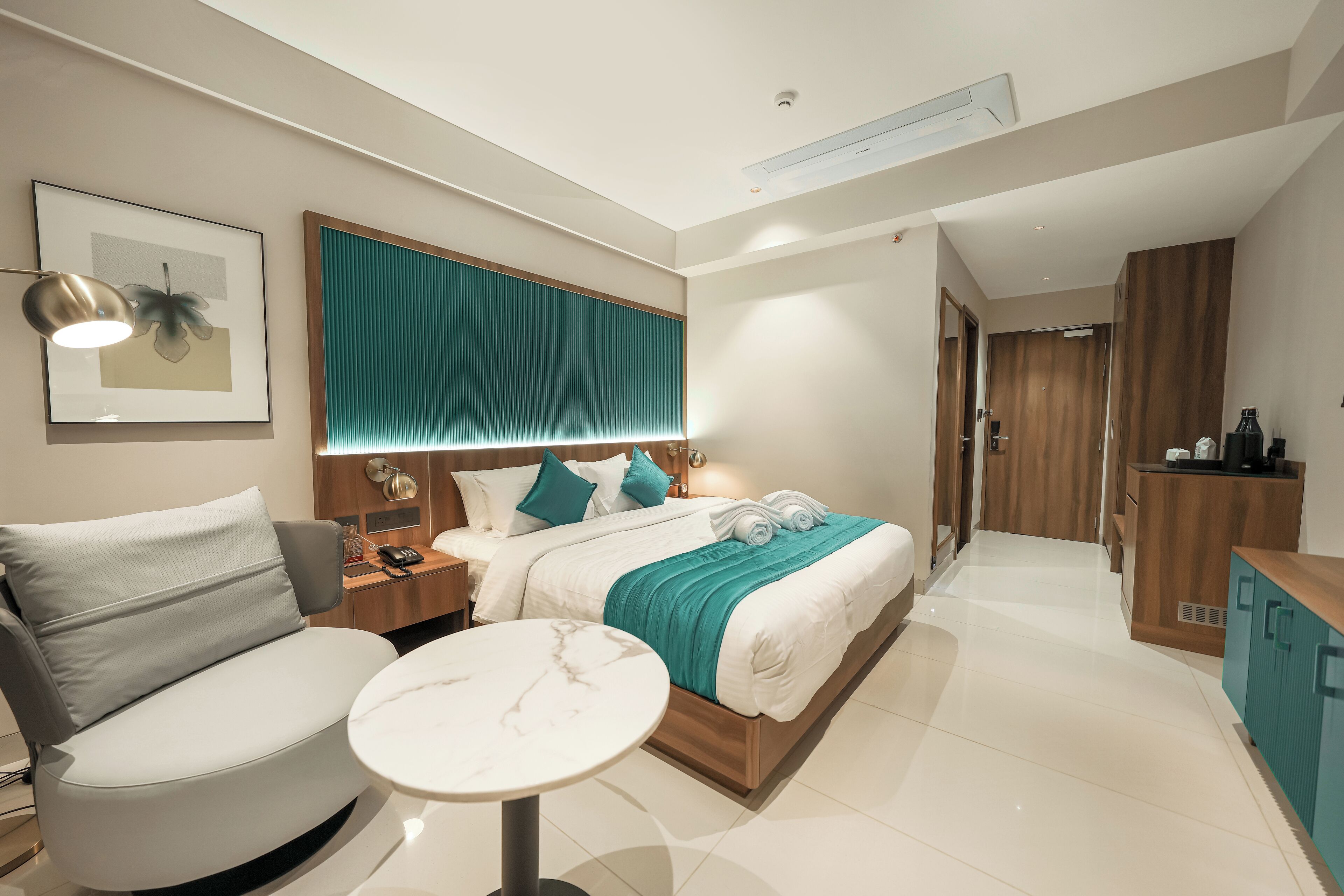 A modern hotel room with a bed, a seating area with a table, a desk, and a wardrobe.