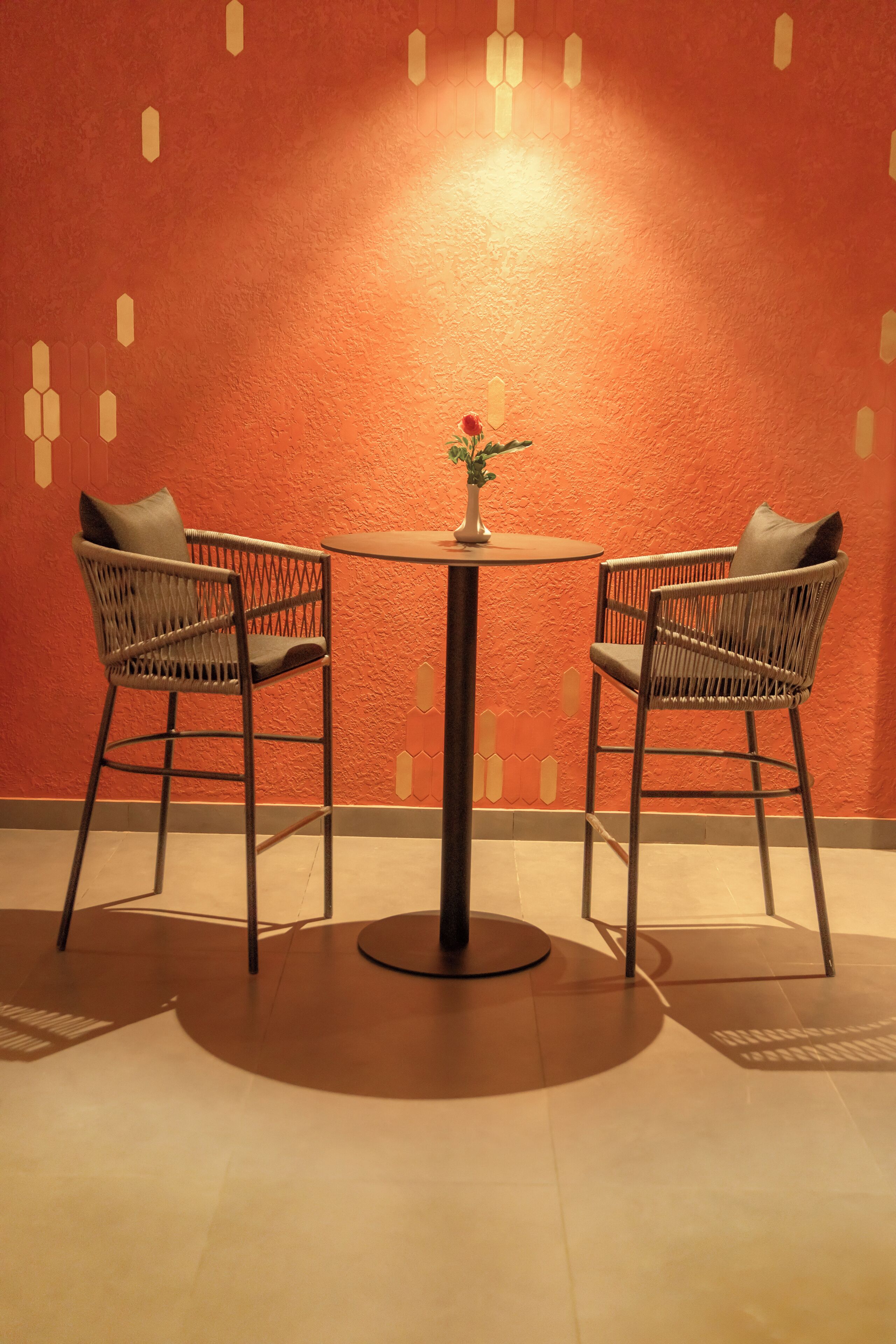 Une table ronde, accompagnée de deux chaises en osier, chacune dotée d’un coussin, est placée contre un mur orange à la texture particulière.