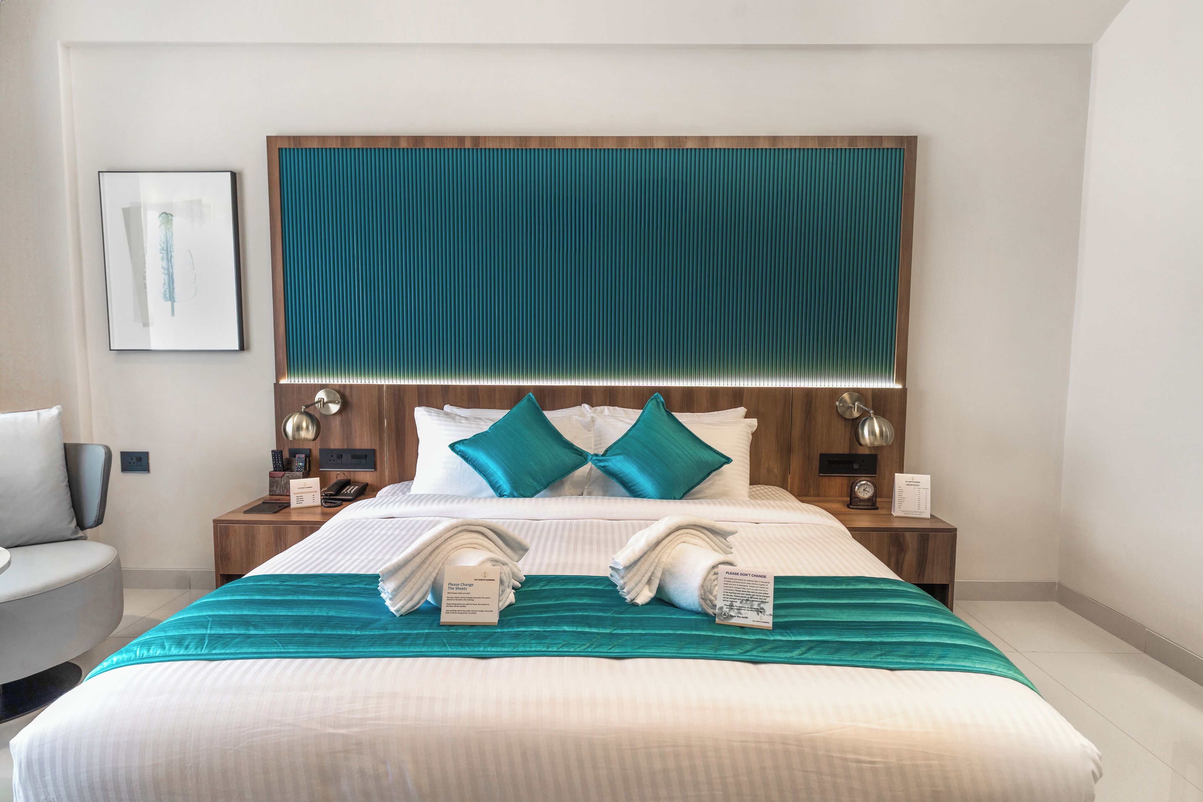 A hotel room with a large bed, two nightstands, a bedside table, a wall-mounted artwork, and a teal-colored headboard.