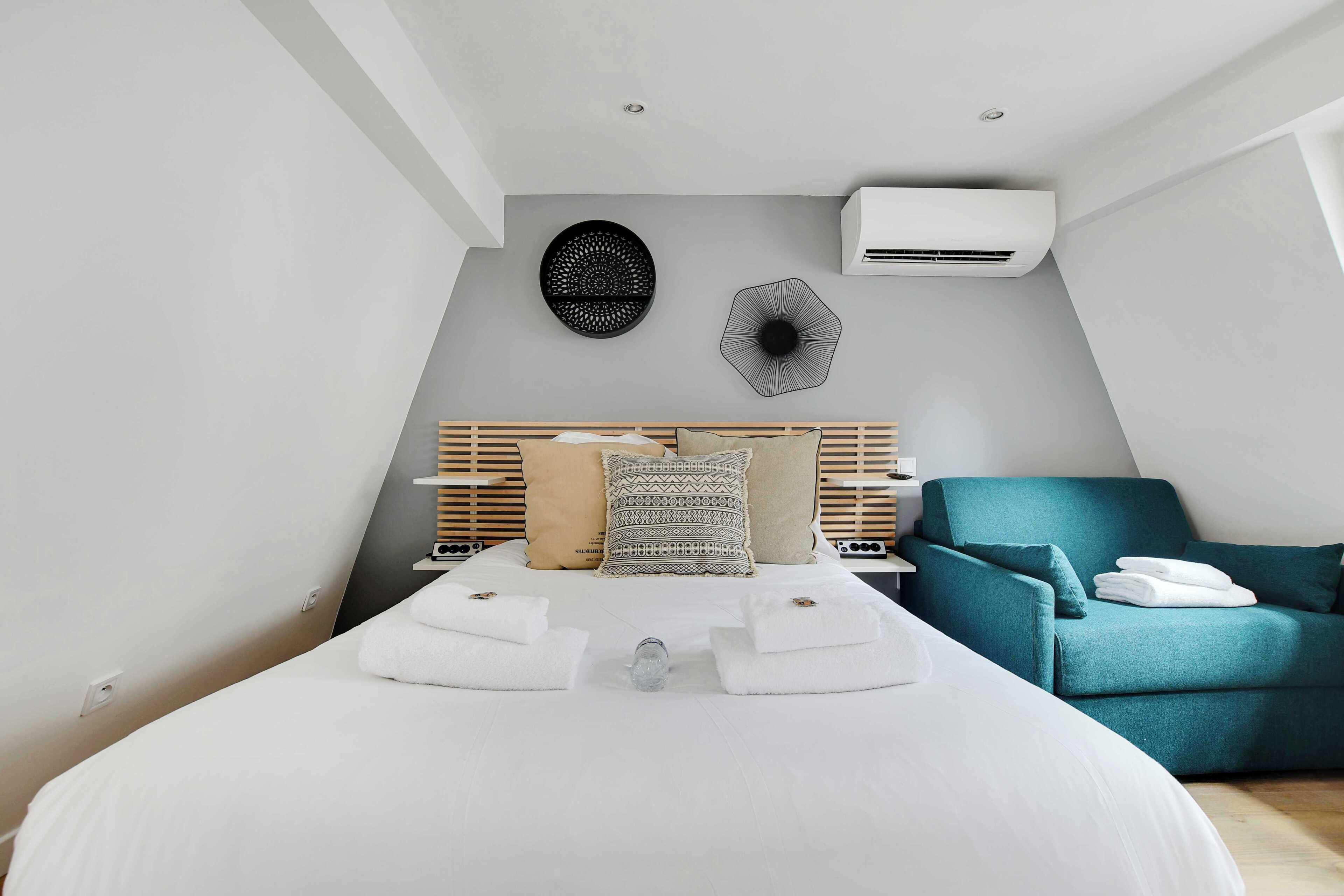 A modern hotel room with a neatly made bed, a teal armchair, and a wall-mounted air conditioner.