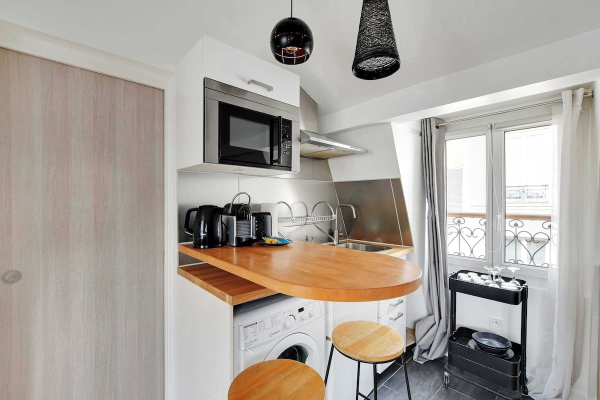 Una cucina compatta con forno a microonde, macchina del caffè e lavatrice. Dispone di un piano di lavoro in legno, due sgabelli e una finestra con vista sul balcone.