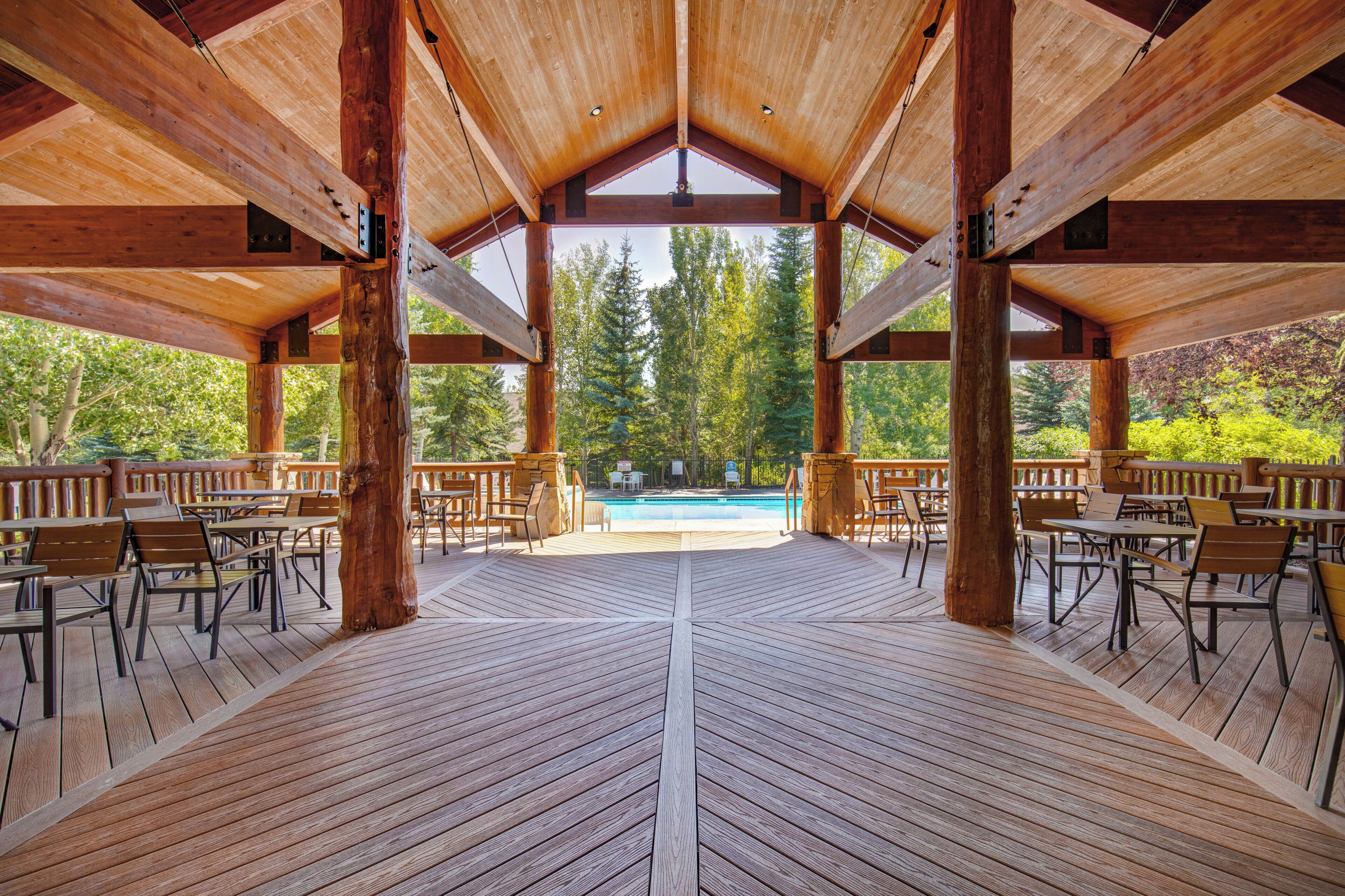 Un espace extérieur abrité, doté d’un plancher en bois, de tables et de chaises, qui offre une vue sur une piscine et est entouré d’arbres.