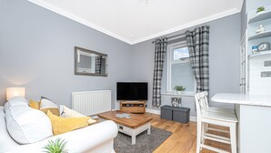 A modern living room with a white sofa, a wooden coffee table, a TV, and a dining area with a white chair and table. - The Wee Haig - 1 Bedroom Condo (Glenrothes)
