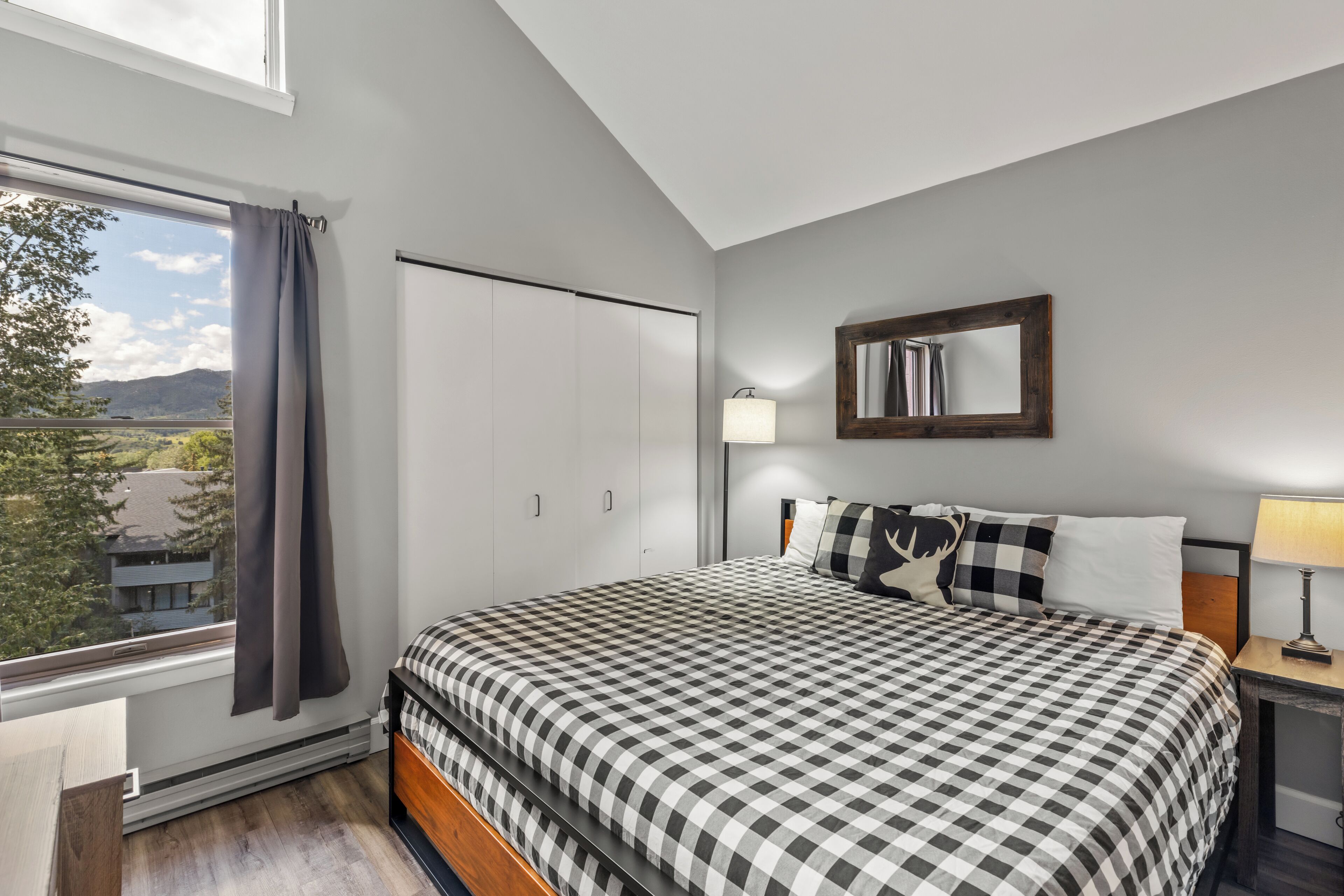 A bedroom with a checkered bedspread, a large window with curtains, a wardrobe, a bedside table with a lamp, and a mirror on the wall.