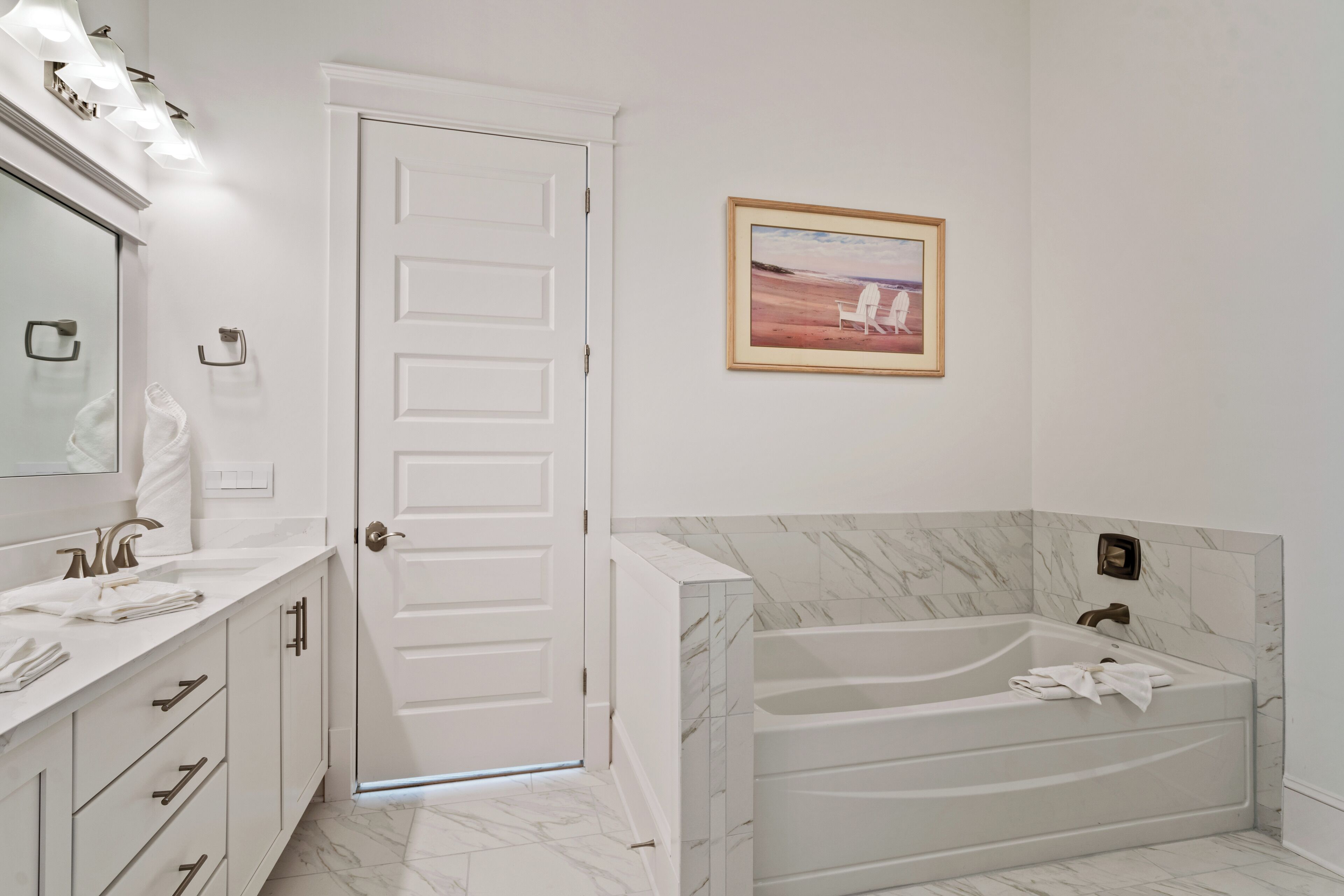 A bathroom with a white door, a large bathtub, a vanity with a mirror and lights, and a framed picture on the wall.