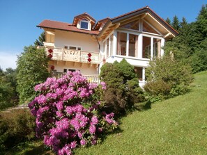 Une maison à deux étages dotée d’un balcon, entourée d’une végétation luxuriante et d’arbustes en fleurs.