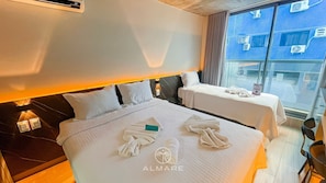 Habitación de hotel con dos camas, una mesita de noche, una lámpara y vistas a la ciudad a través de amplios ventanales.